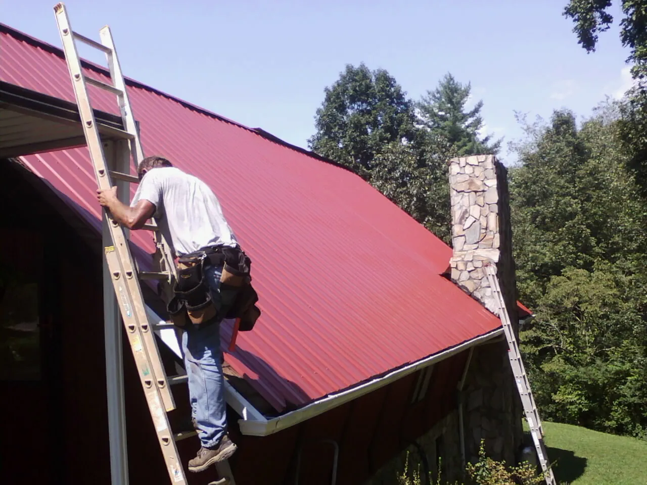 Roofer on a ladder beside a red metal roof and stone chimney — Soffit & Fascia Repair by Roofing Brothers in Blue Island